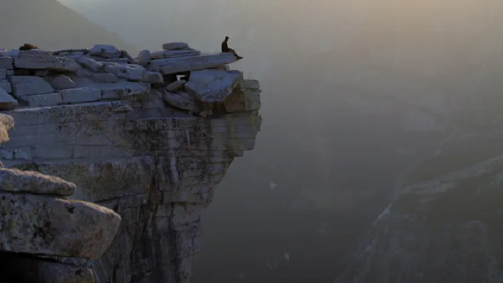 A solitary figure stands on a cliff edge overlooking a vast, hazy canyon at sunrise or sunset.