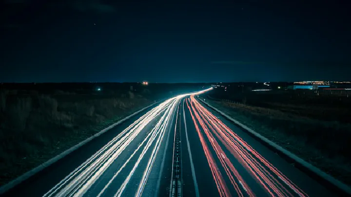 Long exposure photograph of light trails from vehicles moving on a dark highway under a starry night sky.