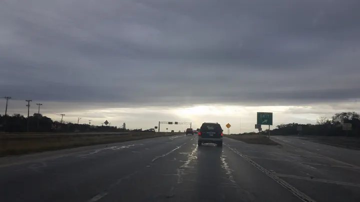 View of a wet highway under stormy clouds, showing a bright sunset glow and an exit sign on the right.