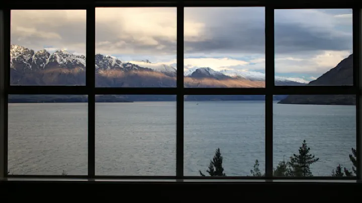View of snowy mountains and a calm bay seen through a large window frame under dramatic, cloudy skies.